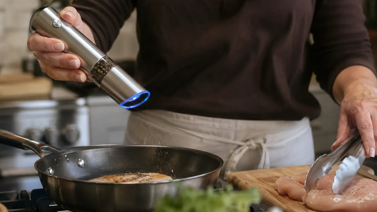 Hand tilting the Circle Joy electric pepper grinder sideways over a pan while the other hand holds tongs over raw chicken