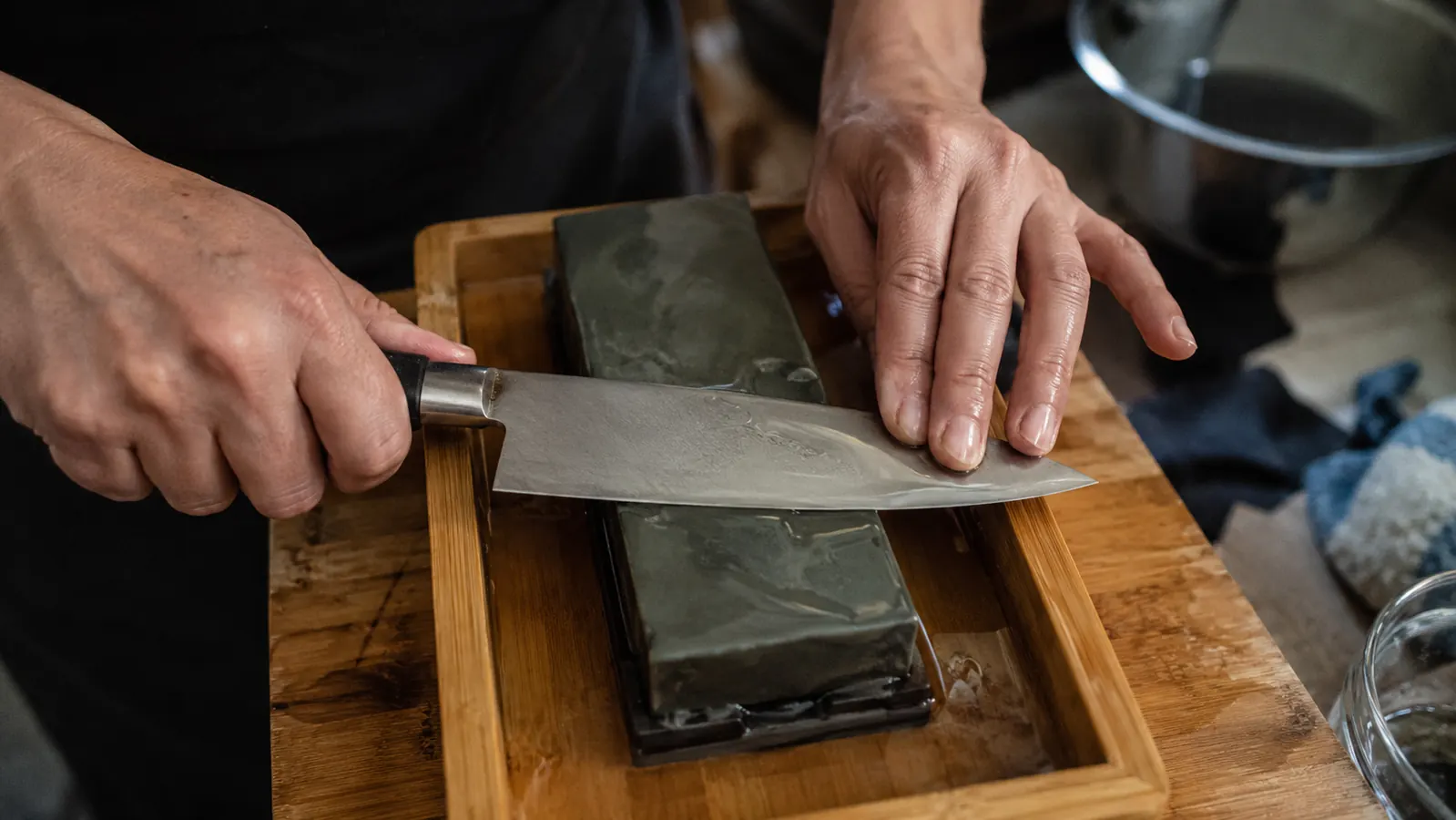 Hands working a chef's knife across a wet whetstone at a low angle, water pooling at the base