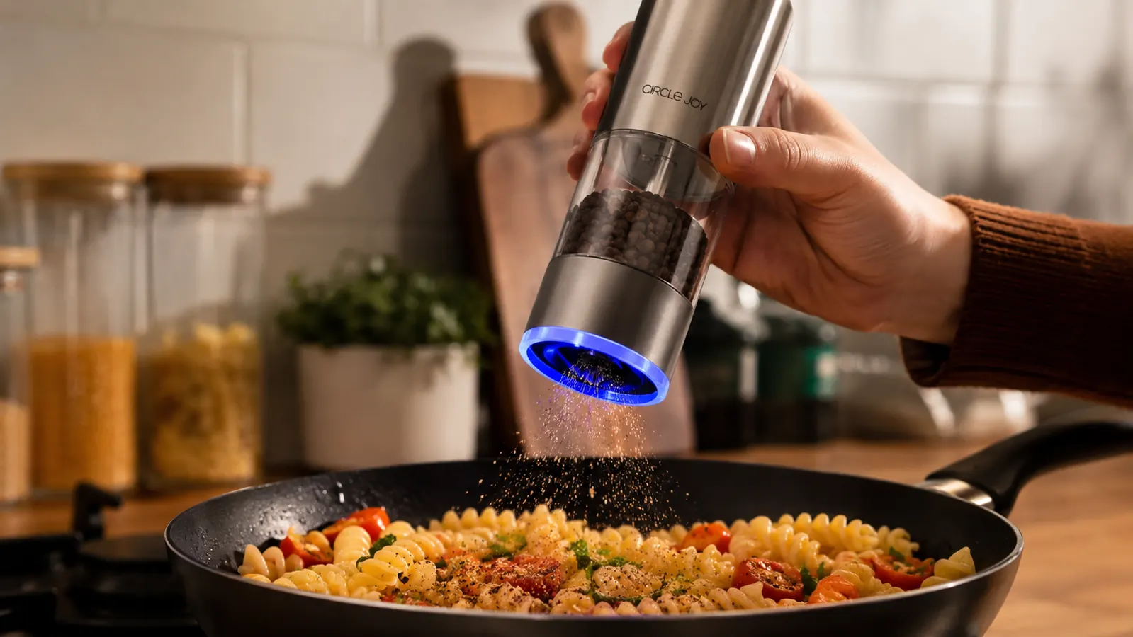 Hand tilting the Circle Joy electric grinder upside down over a pan of pasta, LED light glowing at the base as pepper falls